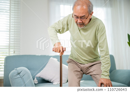 Elderly man suffering from knee pain ache holding handle of cane, senior old man typing to stand up from sofa alone with walking cane stick to walk at home, retirement medical healthcare concept 110726043