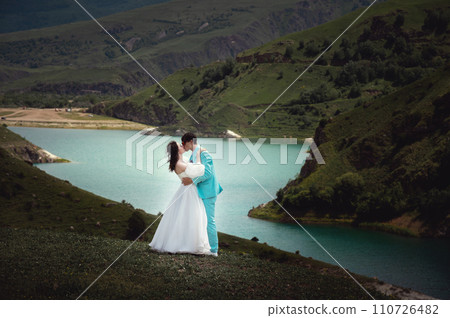Love, wedding and married couple kissing by the lake outdoors in honor of their romantic marriage. Water, summer, mountains or a kiss Love, wedding and married couple kissing by the lake outdoors in honor of their romantic marriage. Water, summer, mountains or a kiss 110726482