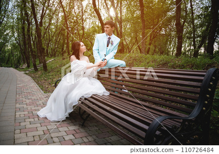European bride and groom are sitting on a park bench. Newlyweds admiring each other while walking through a green summer park European bride and groom are sitting on a park bench. Newlyweds admiring each other while walking through a green summer park 110726484