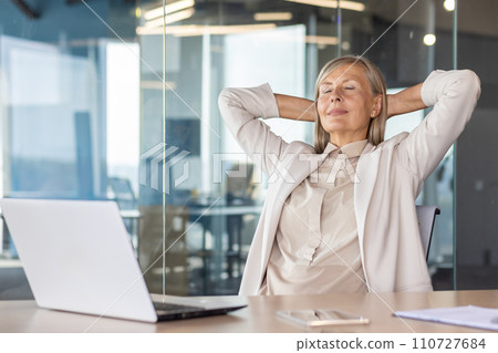 A gray-haired senior business woman is contentedly sitting in the office at the desk with her eyes closed, her hands behind her head and resting. 110727684
