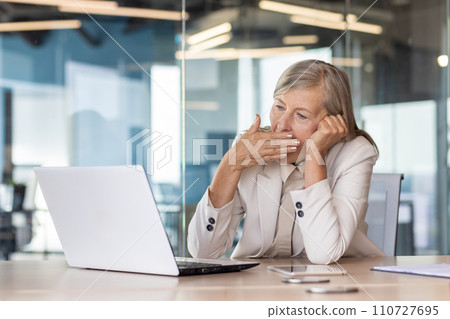 Tired senior woman sitting at desk in front of laptop in office in business suit, head on hand and yawning while covering mouth with hand. 110727695