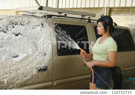 Pretty Asian woman spraying water to wash car which has foam that washes away with water as well. Pretty Asian woman spraying water to wash car which has foam that washes away with water as well. 110732735