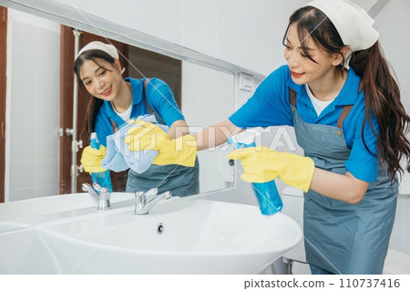 A cheerful maid in rubber gloves wipes bathroom mirror with a rag ensuring cleanliness and shining reflection. Hotel service emphasizes professional cleaning for hygiene. Maid cleaning at home A cheerful maid in rubber gloves wipes bathroom mirror with a rag ensuring cleanliness and shining reflection. Hotel service emphasizes professional cleaning for hygiene. Maid cleaning at home 110737416