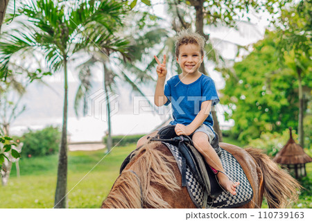 Blond boy riding horse in hotel park on the beach. Sunny summer day happy childhood Blond boy riding horse in hotel park on the beach. Sunny summer day happy childhood 110739163