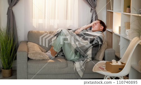 Sickness fever. Seasonal flu. Ill tired man wrapped in blanket laying on couch suffering from bad headache in living room with drugs at home. 110740895
