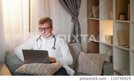 Work leisure. Doctor break. Online communication. Relaxed healthcare specialist man in white uniform typing on laptop sitting on couch in cozy interior. Work leisure. Doctor break. Online communication. Relaxed healthcare specialist man in white uniform typing on laptop sitting on couch in cozy interior. 110740929