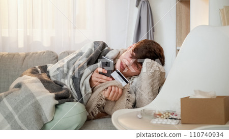 Online pharmacy. Ordering medicine. Internet payment. Tired ill man wrapped in plaid typing smartphone buying with credit card in resting on sofa in home interior. 110740934