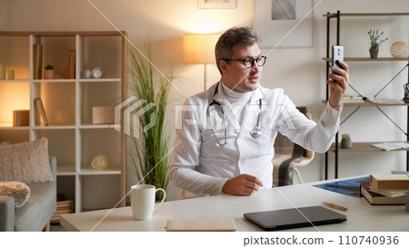 Medical video advice. Health consultation. Online meeting. Healthcare professional man talking at virtual conference on smartphone sitting at desk in clinic interior. 110740936