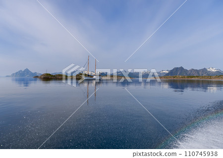 Calm sea mirrors a sailboat and mountains with a faint rainbow in Lofoten Calm sea mirrors a sailboat and mountains with a faint rainbow in Lofoten 110745938