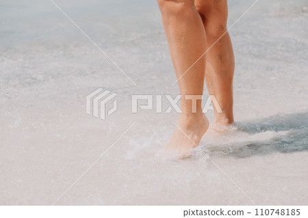 Sea beach travel - woman walking on sand beach leaving footprints in the white sand. Female legs walking along the seaside barefoot, close-up of the tanned legs of a girl coming out of the water. 110748185