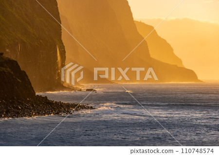 Volcanic rock cliffs Achadas da Cruz in backlit sunlight. Waves of the Atlantic Ocean. Beautiful sunset seascape of the resort island of Madeira, Portugal 110748574