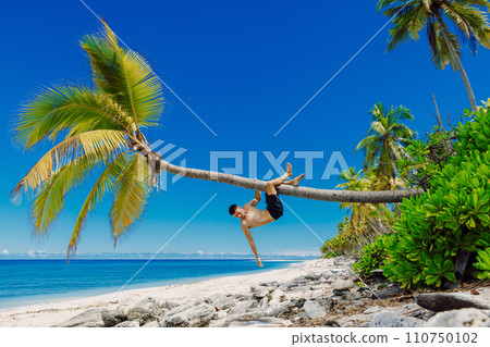 A man shirtless relaxing on tropical beach and climbs a coconut palm tree. Slim man vacation on paradise beach A man shirtless relaxing on tropical beach and climbs a coconut palm tree. Slim man vacation on paradise beach 110750102