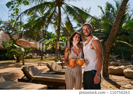 Smiling couple enjoys honeymoon in tropical paradise, sharing fresh coconut water. Fit man and woman in summer attire stand amidst palm trees, celebrating romantic getaway with exotic drinks. Smiling couple enjoys honeymoon in tropical paradise, sharing fresh coconut water. Fit man and woman in summer attire stand amidst palm trees, celebrating romantic getaway with exotic drinks. 110750263