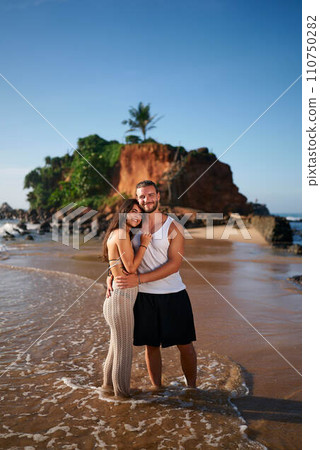 Affectionate couple hugs on sandy beach at sunrise. Romantic getaway by sea, partner embracing by ocean. Love, intimacy by waves, idyllic travel moment, tropical vacation memories. 110750282