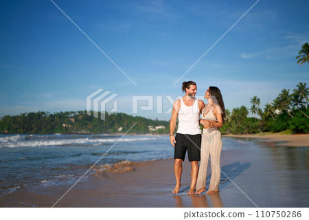Romantic couple strolls on beach, holding hands by the sea. Man, woman enjoy honeymoon at tropical coast. Casual beachwear, sunset walk, love, vacation. Smiling duo in relaxed seaside promenade. Romantic couple strolls on beach, holding hands by the sea. Man, woman enjoy honeymoon at tropical coast. Casual beachwear, sunset walk, love, vacation. Smiling duo in relaxed seaside promenade. 110750286