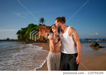 Affectionate couple embrace on tropical beach at sunset. Childfree travelers enjoy serene sea, sandy shore. Young man, woman experience romantic getaway. Love, travel, leisure without kids depicted. Affectionate couple embrace on tropical beach at sunset. Childfree travelers enjoy serene sea, sandy shore. Young man, woman experience romantic getaway. Love, travel, leisure without kids depicted. 110750294