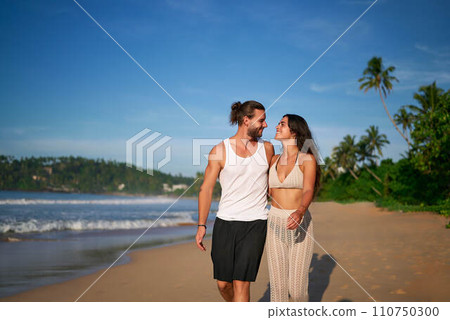 Romantic couple walks on beach at sunset, barefoot man, woman in casual summer clothes enjoy seaside stroll. Loving pair on vacation, leisure travel, coastline wander, tropical getaway, relaxed mood. Romantic couple walks on beach at sunset, barefoot man, woman in casual summer clothes enjoy seaside stroll. Loving pair on vacation, leisure travel, coastline wander, tropical getaway, relaxed mood. 110750300
