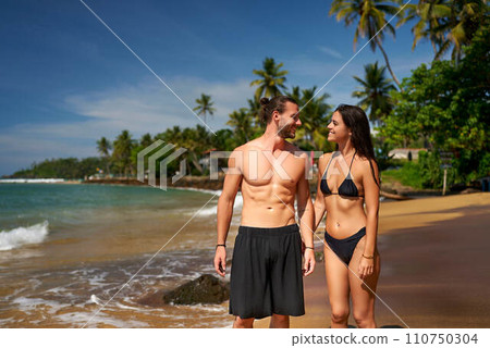 Fit couple enjoys honeymoon on tropical beach. Athletic man and slender woman in swimwear share romantic moment by the sea. Newlyweds celebrate at sunny coastal paradise. Love, travel, intimacy theme. 110750304