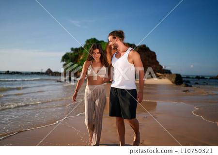 Romantic couple strolls on beach at sunset, holding hands. Woman, man walk barefoot on sand by sea, enjoy vacation. Trendy, young lovers on seaside date. Relaxed, carefree tourists on coastal escape. Romantic couple strolls on beach at sunset, holding hands. Woman, man walk barefoot on sand by sea, enjoy vacation. Trendy, young lovers on seaside date. Relaxed, carefree tourists on coastal escape. 110750321