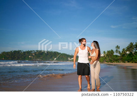 Romantic couple strolls on tropical beach at sunset. Man and woman in love walk barefoot by sea, enjoy honeymoon. Relaxed pair smiles, shares moment on sandy shore. Travel, vacation concept. Romantic couple strolls on tropical beach at sunset. Man and woman in love walk barefoot by sea, enjoy honeymoon. Relaxed pair smiles, shares moment on sandy shore. Travel, vacation concept. 110750322