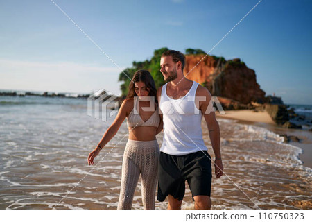 Romantic couple strolls on beach at sunset. Newlyweds enjoy honeymoon by sea, walking barefoot on sand. Man, woman in casual wear share love, laughter. Gentle waves, cliffs backdrop, tranquility. Romantic couple strolls on beach at sunset. Newlyweds enjoy honeymoon by sea, walking barefoot on sand. Man, woman in casual wear share love, laughter. Gentle waves, cliffs backdrop, tranquility. 110750323