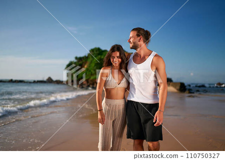 Couple walks on beach, adult duo sans kids enjoys seaside stroll. Childless pair on vacation smile, unwind by ocean, travel trends. Man, woman in summer attire embrace leisure, tropical getaway. Couple walks on beach, adult duo sans kids enjoys seaside stroll. Childless pair on vacation smile, unwind by ocean, travel trends. Man, woman in summer attire embrace leisure, tropical getaway. 110750327