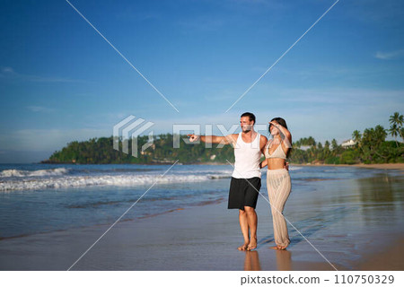 Couple enjoys beach stroll, man pointing horizon, woman smiles, tropical island escape. Ocean sand, vacation vibes, romantic getaway, summer joy coastal walk. Explore relaxing wanderlust paradise. 110750329