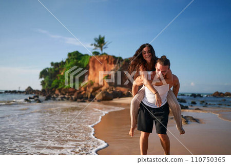 Couple enjoys piggyback ride on tropical beach sunset. Man carries joyful woman on back, strolling along shoreline. Romantic travelers at seaside, exotic holiday escape. Playful duo on sandy coast. 110750365
