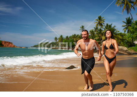 Cheerful man and woman enjoy beach run, barefoot by sea, palm trees background. Couple in active lifestyle, summer vacation sprint. Joyful jog, sunlit coast, ocean living freedom, fitness together. Cheerful man and woman enjoy beach run, barefoot by sea, palm trees background. Couple in active lifestyle, summer vacation sprint. Joyful jog, sunlit coast, ocean living freedom, fitness together. 110750366