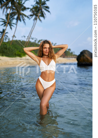 Fit woman in white bikini enjoys sunbathing by tropical beach. Attractive female with healthy tan applies sunscreen for UV protection. Holidays at palm-lined seaside, skin care routine. 110750385