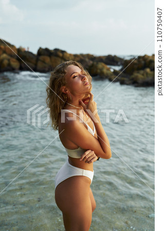 Young woman enjoying sun at seaside, wearing swimsuit, coastal rocks backdrop, serenity concept Young woman enjoying sun at seaside, wearing swimsuit, coastal rocks backdrop, serenity concept 110750407