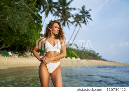 Fit woman enjoys alone time on tropical beach. Solo traveler in swimwear relaxes by sea under palms. Independent female explores sandy coast. Romantic adventure, wellness journey concept. 110750412
