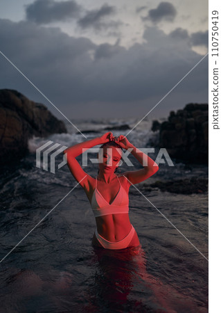 Attractive woman enjoys nocturnal swim in ocean, illuminated by red light. Fashionable female in swimwear relaxing in sea at evening. Nighttime beauty, leisure by beach silhouette. Stylish lady bathes 110750419