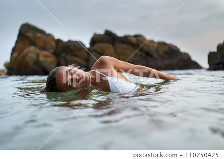 Woman in white swimsuit lies in water near rocky coast. Relaxed female enjoys shallow sea, leisure time at serene beach. Zen moment in nature, tranquil summer escape, ocean relaxation. 110750425
