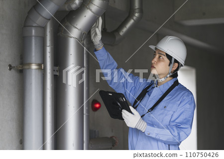 Building maintenance image: A man in work clothes holding a tablet and inspecting piping 110750426