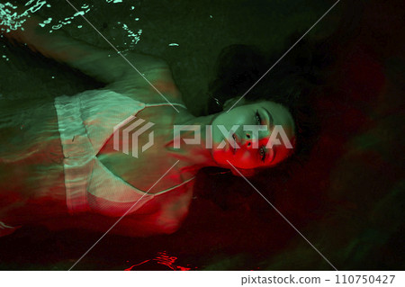 Woman floats in lagoon, red illumination surrounds her, night setting conveys solitude, introspection, eerie atmosphere, concept of inner world exploration and duality of self. 110750427