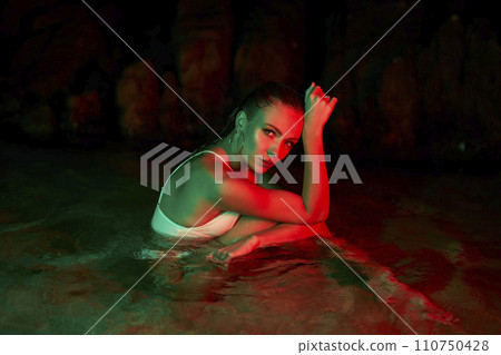 Woman sits in water at night, illuminated by red and green lights, expressing contemplation and moodiness, wet hair, focused gaze, feeling of solitude. 110750428