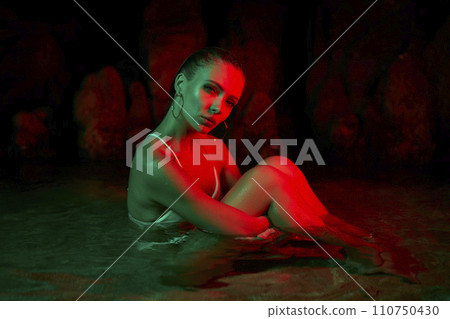 Woman sits in water at night illuminated by green, red lights. Female models in lagoon for creative photography, reflections shimmer on wet skin. Conceptual art shoot, depicts beauty, inner self. 110750430