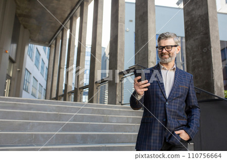 Smiling senior businessman in suit using smartphone while standing on office stairs. Concept of experienced corporate professional. Smiling senior businessman in suit using smartphone while standing on office stairs. Concept of experienced corporate professional. 110756664