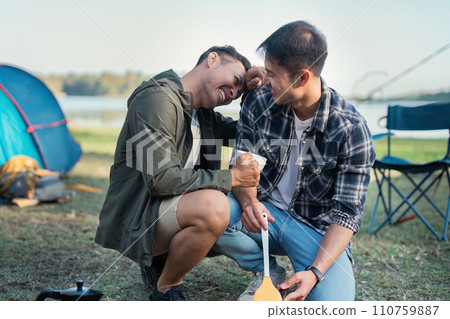 Gay LGBTQIA couple sitting on picnic chair drinking tea and making food while camping on vacation holiday Gay LGBTQIA couple sitting on picnic chair drinking tea and making food while camping on vacation holiday 110759887