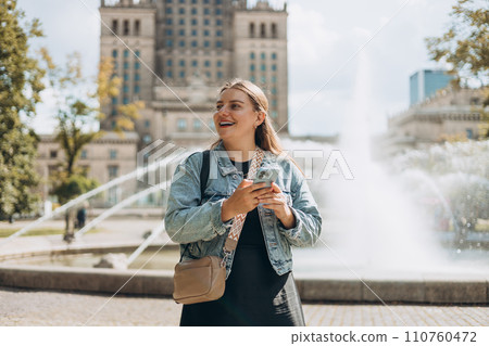 Young tourist woman walking in the park near Palace of Culture and Science in Warsaw city, Poland. Women chatting on cell smartphone while relaxing outdoors. Technology, Urban lifestyle concept 110760472