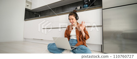 Portrait of happy woman in headphones, looking at laptop, waving hand at computer, chatting on video online, joins team meeting, calling friend on computer, sitting on floor Portrait of happy woman in headphones, looking at laptop, waving hand at computer, chatting on video online, joins team meeting, calling friend on computer, sitting on floor 110760727