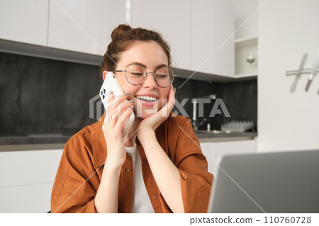 Portrait of beautiful young woman managing her own business from home, freelancer making phone call, sitting in kitchen with laptop and smiling Portrait of beautiful young woman managing her own business from home, freelancer making phone call, sitting in kitchen with laptop and smiling 110760728
