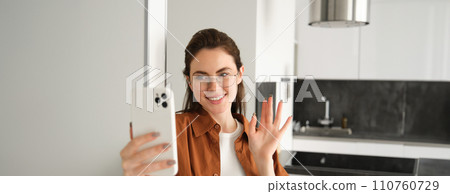 Portrait of stylish beautiful woman at home, video chats on smartphone, connects to online meeting from her living room, lifestyle social media blogger records a video on mobile phone Portrait of stylish beautiful woman at home, video chats on smartphone, connects to online meeting from her living room, lifestyle social media blogger records a video on mobile phone 110760729