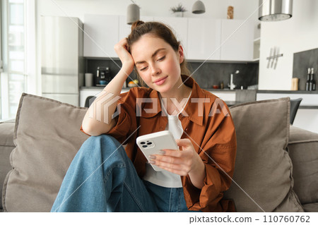 Portrait of young modern woman reading on mobile phone, scrolling social media app on smartphone, sitting on couch in living room in casual clothes Portrait of young modern woman reading on mobile phone, scrolling social media app on smartphone, sitting on couch in living room in casual clothes 110760762