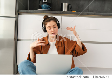 Portrait of woman listens to music in headphones, while working on laptop, sits at home on kitchen floor, making dance movements Portrait of woman listens to music in headphones, while working on laptop, sits at home on kitchen floor, making dance movements 110760765