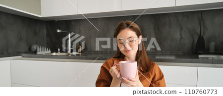 Relaxing day at home. Smiling young woman in glasses, enjoying fresh brewed cup of coffee, holding mug and sitting in kitchen, resting on peaceful autumn day at home Relaxing day at home. Smiling young woman in glasses, enjoying fresh brewed cup of coffee, holding mug and sitting in kitchen, resting on peaceful autumn day at home 110760773