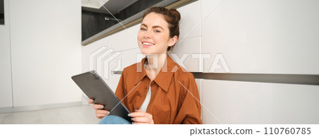 Portrait of young woman working or studying from home, watching her online course on digital tablet, sitting on kitchen floor, smiling and looking happy 110760785