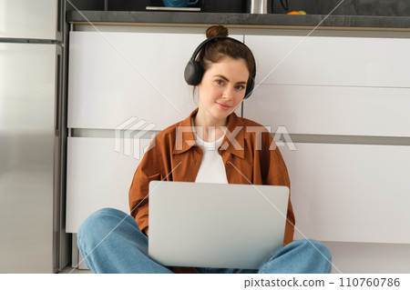 Young woman, student doing homework on laptop, sitting on floor, listening to music in headphones and working from home 110760786