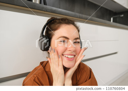 Cute young woman, makes funny faces, listens to music, wears wireless headphones, having fun at home Cute young woman, makes funny faces, listens to music, wears wireless headphones, having fun at home 110760804